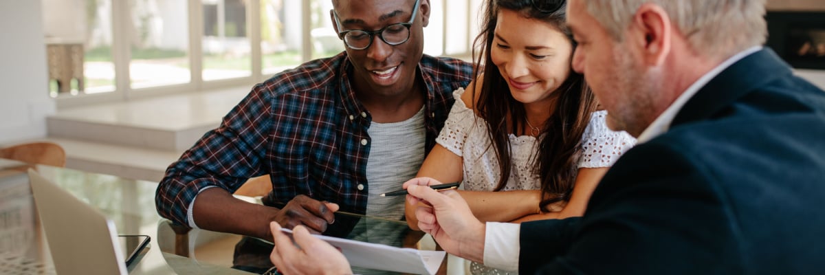 Male real estate agent offering to buy a new house to young couple during a meeting. Realtor presenting technical documentation to owner couple.